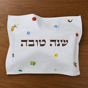 Puede incluir: Un paño rectangular blanco con el texto hebreo marrón "שנה טובה" en el centro. La tela está decorada con pequeños iconos bordados que incluyen un tarro de miel, una abeja, una estrella de David, una granada, un pájaro, un shofar, una manzana y flores.