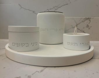 May include: A white ceramic set featuring three pieces: a small round candle holder with a lit candle, a cylindrical container, and a round lidded jar. The set is arranged on a matching white circular tray. Hebrew text is etched on each piece.