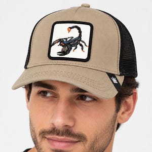 Pode incluir: Um boné de beisebol bege com a parte traseira em rede preta e um emblema quadrado na frente. O emblema apresenta uma ilustração detalhada de um escorpião preto com detalhes em laranja sobre um fundo branco. Uma pequena etiqueta preta com um logótipo de coroa branca encontra-se na pala.