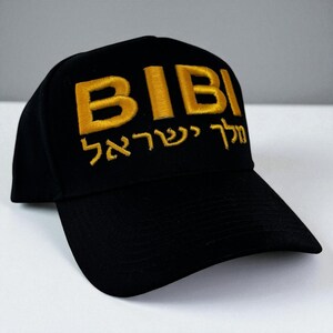 May include: Black baseball cap with the embroidered word "BIBI" in gold, above Hebrew text. The cap has a curved brim and a structured crown, suitable for casual wear.