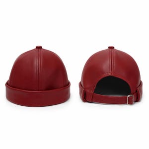 May include: Two views of a deep red, brimless cap made of a smooth, leather-like material. The cap features a rounded crown with stitched panels, a wide folded cuff, and an adjustable strap with a silver-toned metal buckle at the back.