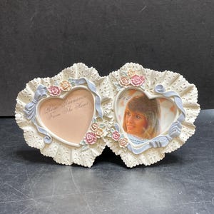 May include: Cream-coloured double heart-shaped photo frame with floral and ribbon accents. The frame has a scalloped edge and holds two 5 x 5 cm photos. Text on the frame reads "Rose Romance From The Heart."