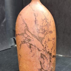 May include: A terracotta-coloured ceramic vase with a narrow neck and rounded body. Dark brown, vein-like patterns cover the surface, creating a marbled effect. The vase has a smooth, matte finish and a slightly off-white rim.