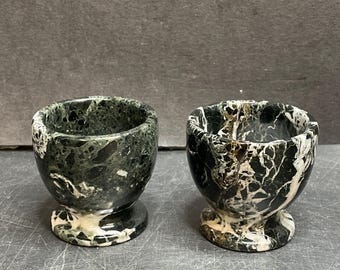 May include: Two marble egg cups. One is primarily dark green with white veining, the other is black and white. Both have a rounded bowl shape and sit on a pedestal base. The cups are made of a natural stone material.