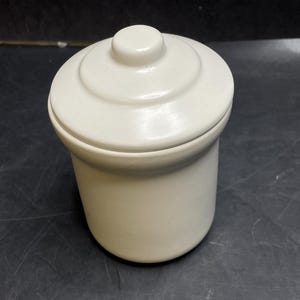 May include: A cream-coloured ceramic jar with a lid. The jar is cylindrical with a rounded base and a lid with a small, circular knob. The surface of the jar and lid is smooth and has a matte finish. The jar is sitting on a dark surface.