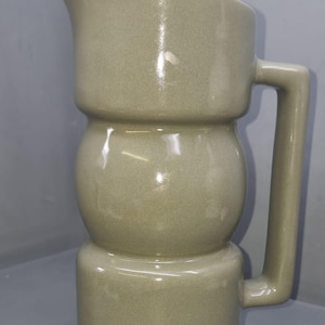 May include: A sage green ceramic jug with a unique, bulbous design. The jug features a curved spout and a rectangular handle. The overall shape is a combination of cylindrical and spherical forms, creating a distinctive aesthetic. The jug is approximately 25 cm tall.