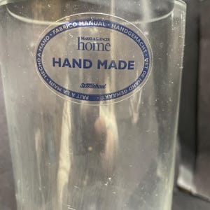May include: A clear, cylindrical glass with a blue and white label that reads "HAND MADE" and "home" from Marks & Spencer. The label also includes text in multiple languages, including "FABRICO MANUAL" and "FAIT A LA MAIN".