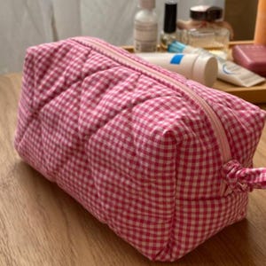 Op de afbeelding: Een rechthoekige gewatteerde toilettas met een roze en wit vichy-ruitpatroon. De tas heeft een ritssluiting aan de bovenkant met een bijpassend stoffen treklipje. De gewatteerde textuur creëert een ruitpatroon over het buitenoppervlak.