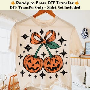 May include: Cream-colored t-shirt featuring a Halloween-themed design. The graphic includes two orange jack-o'-lanterns with green stems and a bow, surrounded by black star shapes. Text at the top reads "Ready to Press DTF Transfer".