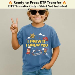 以下が含まれることがあります： 青いTシャツで、I KNEW IT I KNEW YOUというテキスト、黄色い保安官の星、赤いカウボーイハット、赤いカウボーイブーツ、ビーズのブレスレット、積み重ねられたアルファベットブロックのグラフィックプリントが施されており、小さな白い雲が周囲に描かれています。