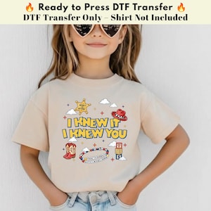 以下が含まれることがあります： I KNEW IT I KNEW YOUというテキストがプリントされたベージュのTシャツ。デザインには、黄色の保安官バッジ、赤いカウボーイハット、赤いカウボーイブーツ、ビーズのブレスレット、TとSの文字が書かれた積み木が含まれています。