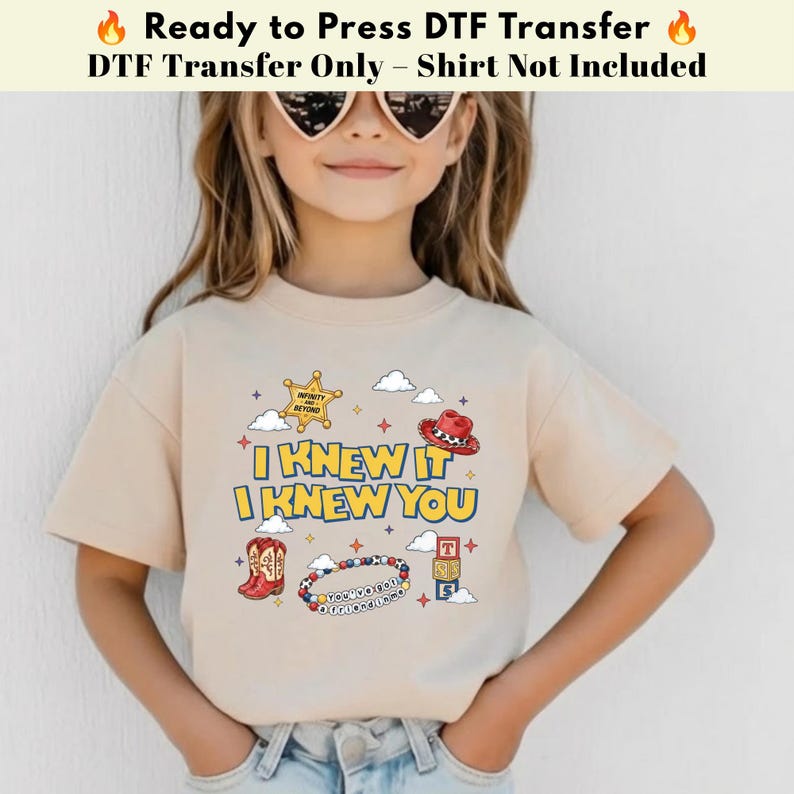 以下が含まれることがあります： I KNEW IT I KNEW YOUというテキストがプリントされたベージュのTシャツ。デザインには、黄色の保安官バッジ、赤いカウボーイハット、赤いカウボーイブーツ、ビーズのブレスレット、TとSの文字が書かれた積み木が含まれています。
