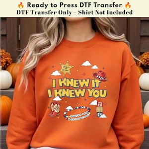 以下が含まれることがあります： I KNEW IT I KNEW YOUというテキスト、黄色い保安官バッジ、赤いカウボーイハット、赤と白のカウボーイブーツ、ビーズのブレスレット、3つのカラフルなアルファベットブロックが描かれたグラフィックプリント付きのオレンジ色のスウェットシャツ。