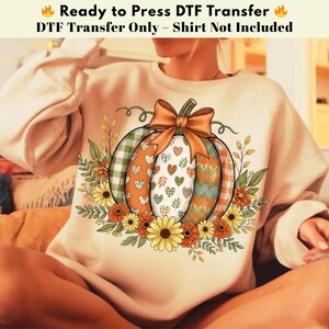 Peut inclure: Un sweat-shirt beige avec un motif de citrouille orné d'un nœud, de cœurs et de motifs floraux. La citrouille présente des sections à carreaux, à cœurs et à chevrons. Le texte en haut indique "Ready to Press DTF Transfer - Shirt Not Included."