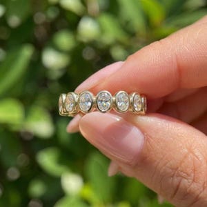 以下が含まれることがあります： 楕円形のクリアな宝石が連なったゴールドバンドリング。ぼやけた緑色の背景を背景にリングが保持され、リングのデザインが際立っています。このリングはジュエリーです。