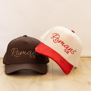 May include: Two baseball caps. One is brown with the word "Romans 5:8" embroidered in a lighter brown. The other is white with a red brim and the same text embroidered in red. Both caps have a curved brim and a button on top.