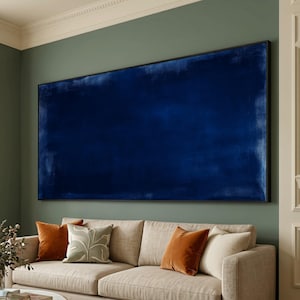 May include: A large, rectangular abstract painting in shades of deep blue, mounted on a wall above a beige sofa. The painting has a dark frame and a textured appearance. Accent pillows in orange and green hues complement the room's decor.