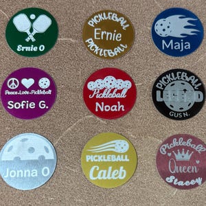 Puede incluir: Una colección de nueve fichas circulares con temática de pickleball. Cada ficha presenta un diseño único, que incluye palas de pickleball, una pelota de pickleball con estela y texto como "Ernie O", "Maja" y "Pickleball Legend". Las fichas son de varios colores.