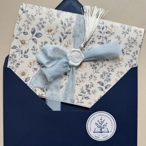Könnte beinhalten: Ein dunkelblauer Umschlag mit einer cremefarbenen Karte mit blauem Blumenmuster. Die Karte ist mit einem hellblauen, ausgefransten Band, einem weißen Wachssiegel mit Blumenmotiv und einer weißen Quaste versehen. Ein runder Aufkleber trägt die Aufschrift The Literary Correspondence Club.