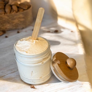 Puede incluir: Un tarro de cristal transparente lleno de crema beige claro, con una cuchara de madera insertada. La tapa de madera del tarro está colocada al lado. La crema tiene una textura suave y batida. Una cesta de nueces es visible en el fondo.