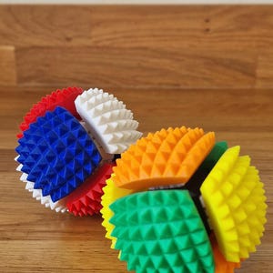 May include: Two spherical sensory toys featuring a textured surface with small, pointed spikes. One toy is composed of red, white, and blue segments, while the other toy is composed of orange, green, and yellow segments, each with a central core.