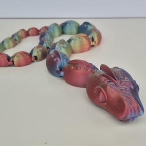 May include: A multi-coloured articulated dragon toy in shades of red, blue, and green. The dragon is made of connected pieces, with a detailed head and body. The toy is designed to be flexible and poseable.