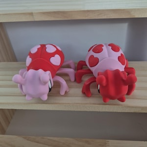 May include: Two mushroom-shaped figurines. One has a red cap with pink hearts and a pink body, the other has a pink cap with red hearts and a red body. Both have pink legs and arms.