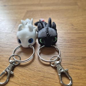 May include: Two dragon-shaped keyrings, one white and one black, each with a silver keyring and clasp. The white dragon has blue eyes, and the black dragon has green eyes. The keyrings are on a wooden surface.