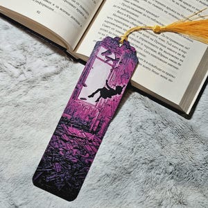 May include: A rectangular bookmark featuring a vibrant purple and pink gradient background with a black silhouette of a person falling through a window. The bookmark has a yellow tassel attached to the top and is resting on an open book with text.