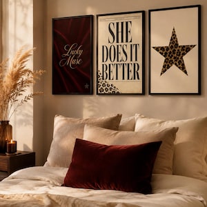 Peut inclure: Trois affiches encadrées. L'affiche de gauche est rouge foncé avec le texte "Lucky Muse". L'affiche centrale est crème avec le texte "SHE DOES IT BETTER" et un motif léopard. L'affiche de droite est crème avec une grande étoile léopard.