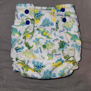 May include: White cloth diaper with a colorful dinosaur print. The diaper features various dinosaurs in shades of green and blue, with the word "Rowr" scattered throughout. It has snap closures in blue and green.