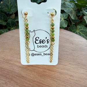 Puede incluir: Pendientes de cadena dorada con cuentas verdes y doradas. Los pendientes se muestran en una tarjeta blanca con el texto "Ese's beads" y el identificador de Instagram "@eses_beads". El fondo presenta follaje verde.