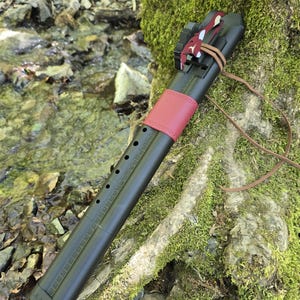 May include: A black Native American flute with a red leather band and a decorative piece at the top. The flute rests on a moss-covered tree trunk near a stream. A leather cord is attached.