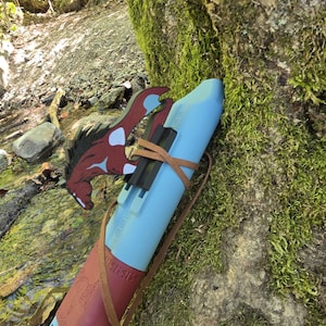 May include: A light blue Native American style flute with a brown leather wrap and a decorative horse head. The flute is resting against a moss-covered tree in a natural outdoor setting.