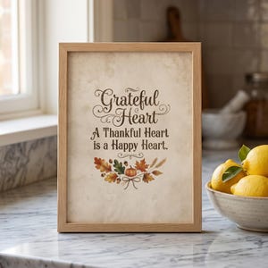 Può includere: Una cornice rettangolare in legno chiaro contenente una stampa color crema con il testo "Grateful Heart A Thankful Heart is a Happy Heart." Il design presenta un'illustrazione decorativa di foglie autunnali, una piccola zucca e spighe di grano.
