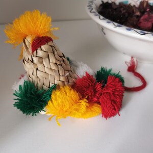 May include: A decorative rooster figurine with a woven straw body, yellow yarn crest, and colorful pom-pom accents in green, red, and yellow. The rooster has a red yarn tail and is placed on a white surface.