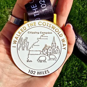 May include: A white and gold medal with the text "I WALKED THE COTSWOLD WAY" and "102 MILES". The medal features a map and illustrations of people, buildings, and sheep. A black ribbon with white text is attached.