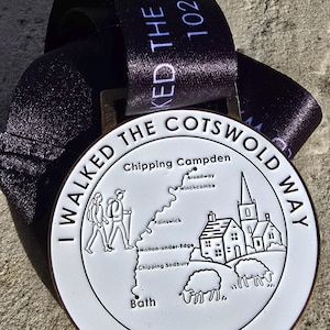 Może przedstawiać: Biało-srebrny medal z czarną wstążką. Medal przedstawia mapę z miastami i napisem "I WALKED THE COTSWOLD WAY 102 MILES". Wstążka ma napis "WALKED THE COTSWOLD WAY 102M".
