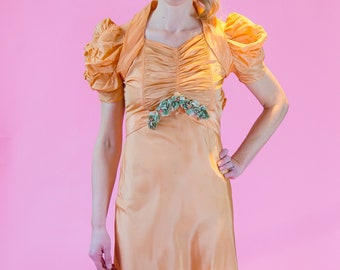 May include: A woman wearing a long, peach-colored dress with a ruffled hem and puffed sleeves. The dress has a green floral embellishment at the waist. She is wearing green heeled sandals.