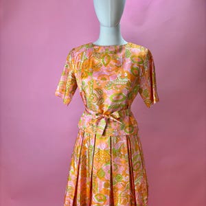 May include: A vintage two-piece dress set on a mannequin. The set features a short-sleeved top and a pleated skirt, both in a vibrant paisley pattern of orange, pink, and green. A matching belt cinches the waist. The dress is a retro style.