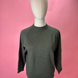 May include: A dark green sweater with a mock neck and long sleeves. The sweater is displayed on a mannequin against a pink background. The sleeves are raglan style.