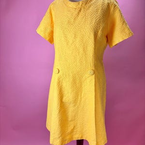 May include: A bright yellow, short-sleeved dress with a textured pattern and two large buttons. The dress is displayed on a mannequin against a pink background. The dress has a round neckline and a slightly A-line shape.