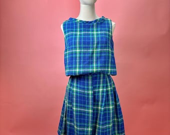 May include: A vintage two-piece outfit on a mannequin. The set includes a sleeveless top and a flared skirt, both in a blue, green, and white plaid pattern. The outfit is displayed against a pink backdrop.