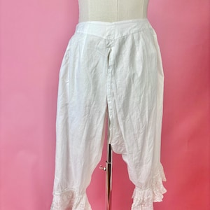 May include: White cotton bloomers featuring a high-waisted design with a split front opening. The leg openings are finished with delicate white lace trim and ruffled edges, providing a classic look for these vintage-style undergarments.