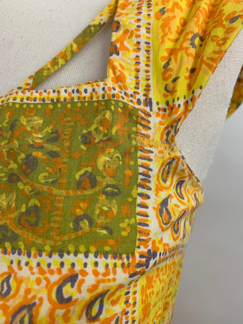 May include: Close-up of a yellow, orange, and green patterned fabric with a paisley design. The fabric has a cut-out detail at the neckline.