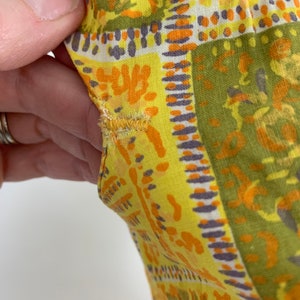 May include: Close-up of a yellow fabric with a geometric pattern in shades of orange, green, and purple. The fabric has a slightly distressed look, with some fraying along the edges.