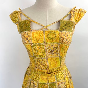 May include: A yellow and green patterned dress with a criss-cross strap detail at the neckline. The dress has a fitted bodice and a full skirt.