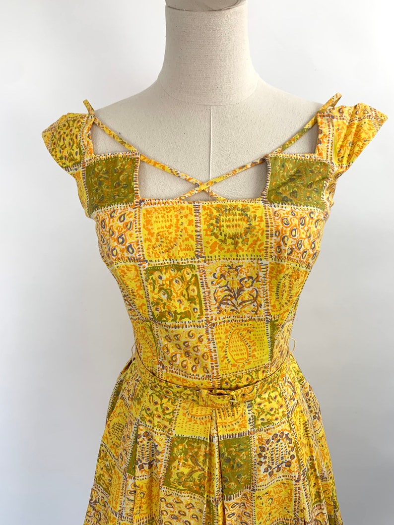 May include: A yellow and green patterned dress with a criss-cross strap detail at the neckline. The dress has a fitted bodice and a full skirt.