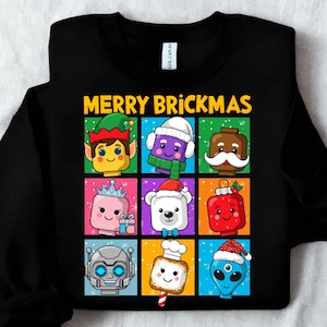 May include: Black sweatshirt with a colorful graphic that says "MERRY BRICKMAS". The graphic features nine cartoon characters in a grid, each resembling a Lego brick figure with festive holiday accessories like elf hats and Santa hats.