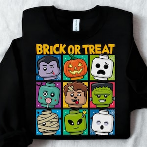May include: Black sweatshirt with the text "BRICK OR TREAT" in yellow. The design features nine colourful squares with Halloween-themed Lego-style characters, including a vampire, pumpkin, and Frankenstein's monster.